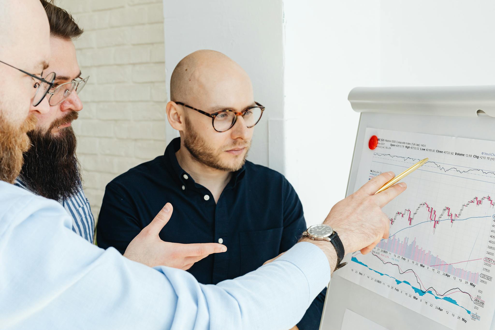 Three men discussing financial charts on a whiteboard during a business meeting.